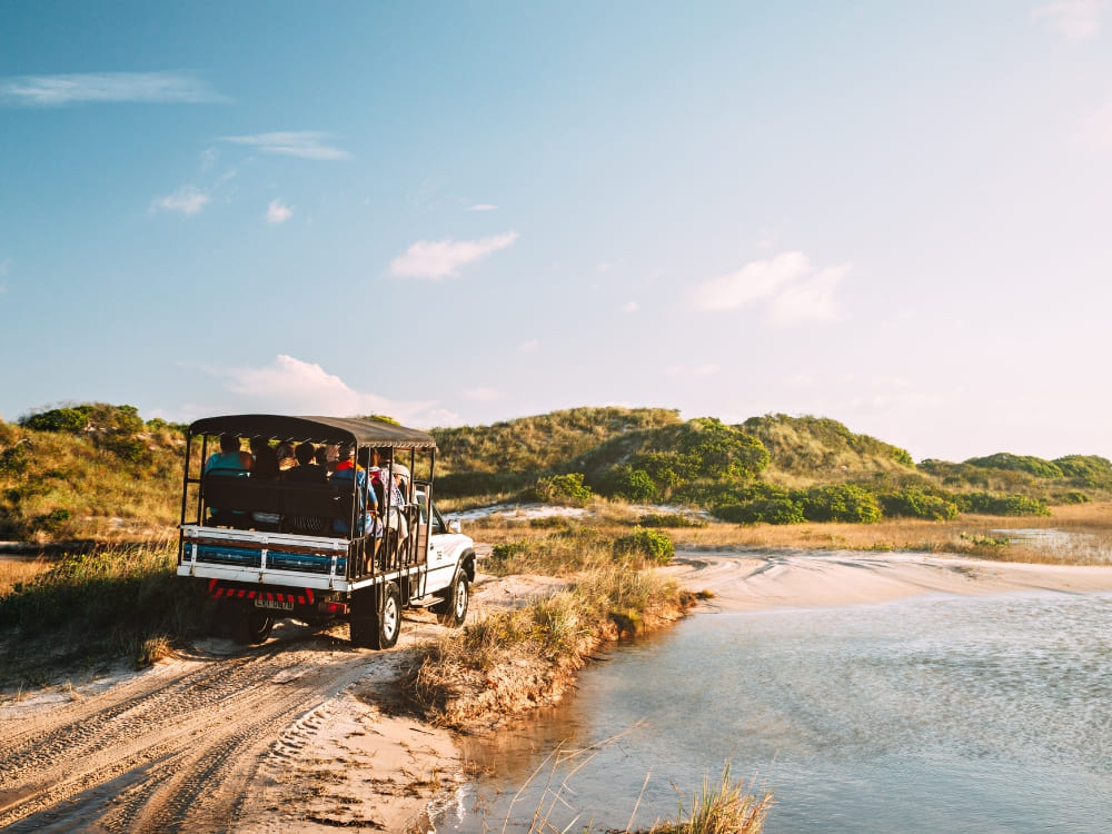 Des touristes dans un véhicule tout-terrain adapté roulent sur une piste inondée et sablonneuse, illustrant les options de transport pour explorer les lagunes des Lençóis Maranhenses.