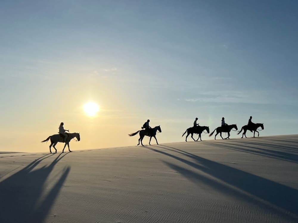 Silhouettes de cinq personnes montant à cheval à travers les dunes de sable des Lençóis Maranhenses sous le soleil éclatant de la fin d'après-midi.