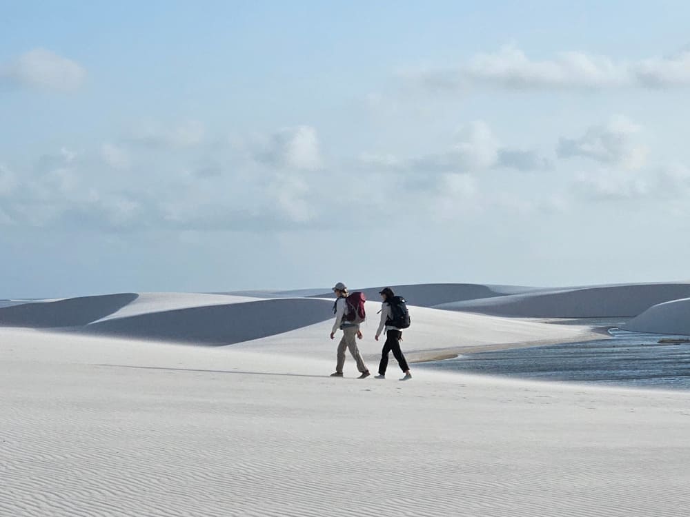 Deux voyageurs avec sac à dos marchent sur les vastes dunes de sable blanc à côté d'une lagune d'eau douce dans le parc national des Lençóis Maranhenses.