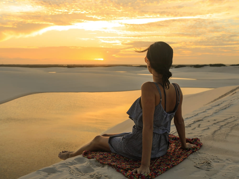 Femme assise sur un paréo de plage sur le sable blanc, admirant le coucher de soleil doré se reflétant sur les eaux des lagunes des Lençóis Maranhenses.