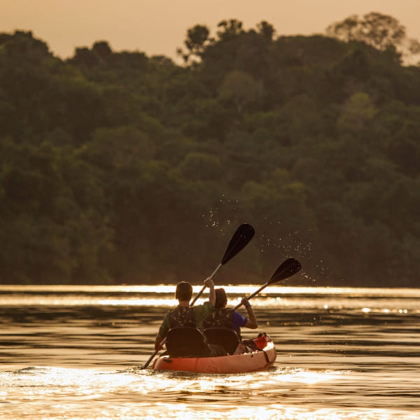 Twee mensen peddelen in een kano op een rustige rivier waarin de zonsondergang in de Amazone weerspiegelt.