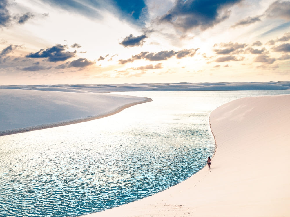 Une personne solitaire marche au bord d'un lagon d'un bleu éclatant qui serpente à travers de vastes dunes de sable blanc sous un ciel nuageux spectaculaire.