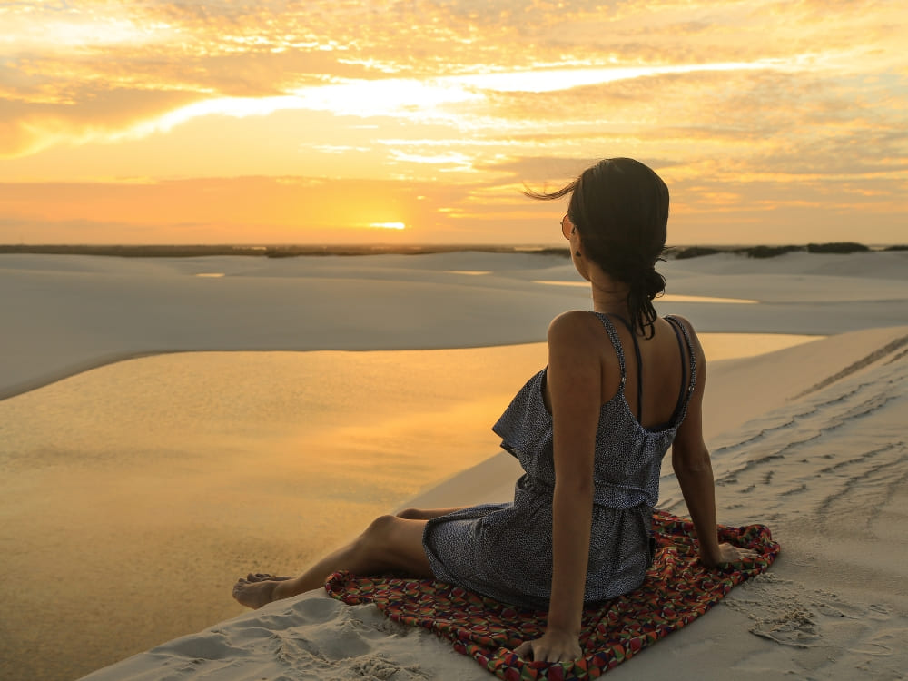 Une femme assise sur une serviette à motifs au sommet d’une dune profite de la chaleur et observe le coucher de soleil au-dessus d’une lagune paisible dans le Parc national des Lençóis Maranhenses.