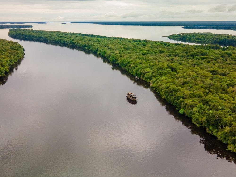 An aerial view shows a small boat traveling down a wide, dark river bordered by dense green tropical forest.