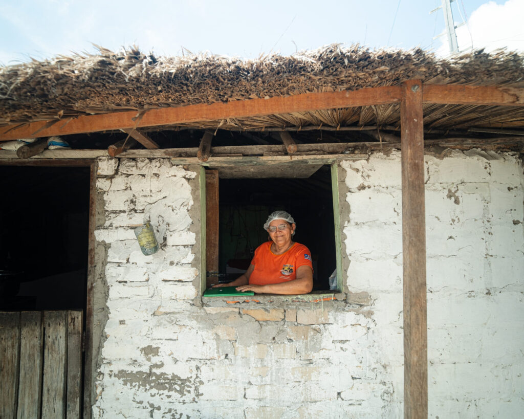 Mulher sorrindo na janela de uma casa rústica de pau a pique. Ela usa blusa laranja e touca, representando as anfitriãs e mulheres do ecoturismo.