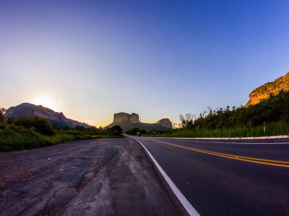An empty paved highway leads toward distant, prominent flat-topped mountains under a bright, setting sun.