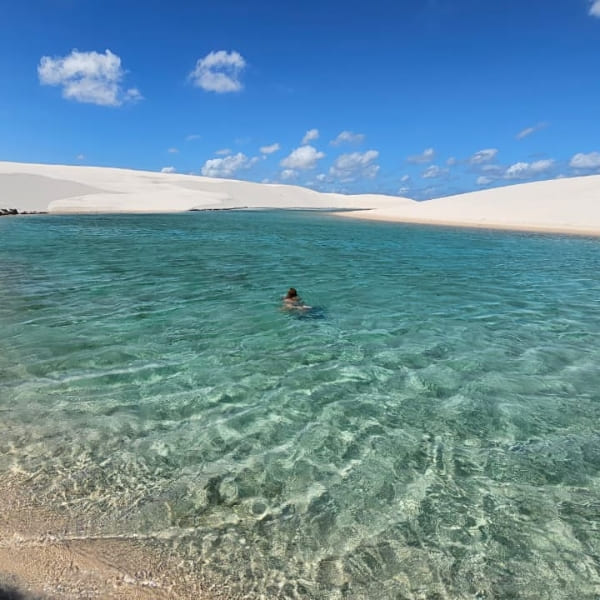 Une personne nage dans une vaste lagune aux eaux bleues transparentes, entre les grandes dunes de sable des Lençóis Maranhenses.