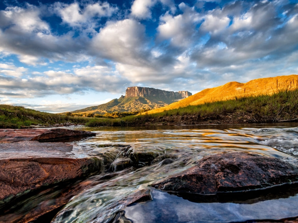 A shallow stream flows over smooth rocks in the foreground, with a grassy plain and a flat-topped mountain bathed in golden light behind it.