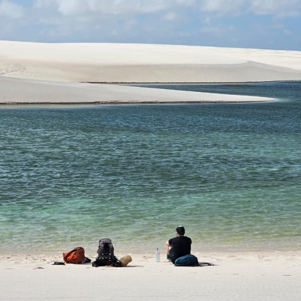 Un randonneur se repose sur le sable blanc, contemplant les eaux calmes et vertes d'une lagune aux Lençóis Maranhenses.