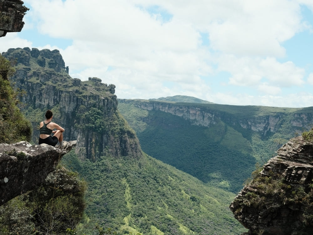 A woman sits on the edge of a steep rocky cliff, taking in the view of a massive, green canyon and distant mountains.
