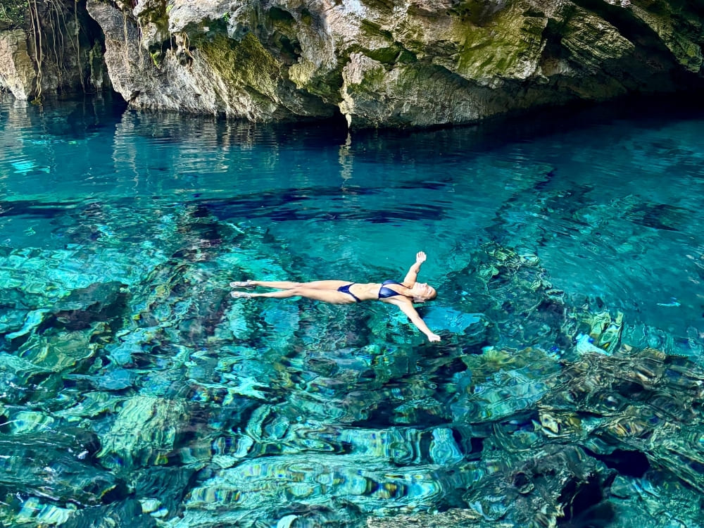 A woman in a dark swimsuit floats peacefully on her back in crystal-clear, bright blue water near a rocky cave wall.