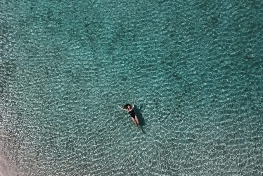 An aerial view shows a woman in a dark swimsuit floating peacefully on her back in clear, bright turquoise water.