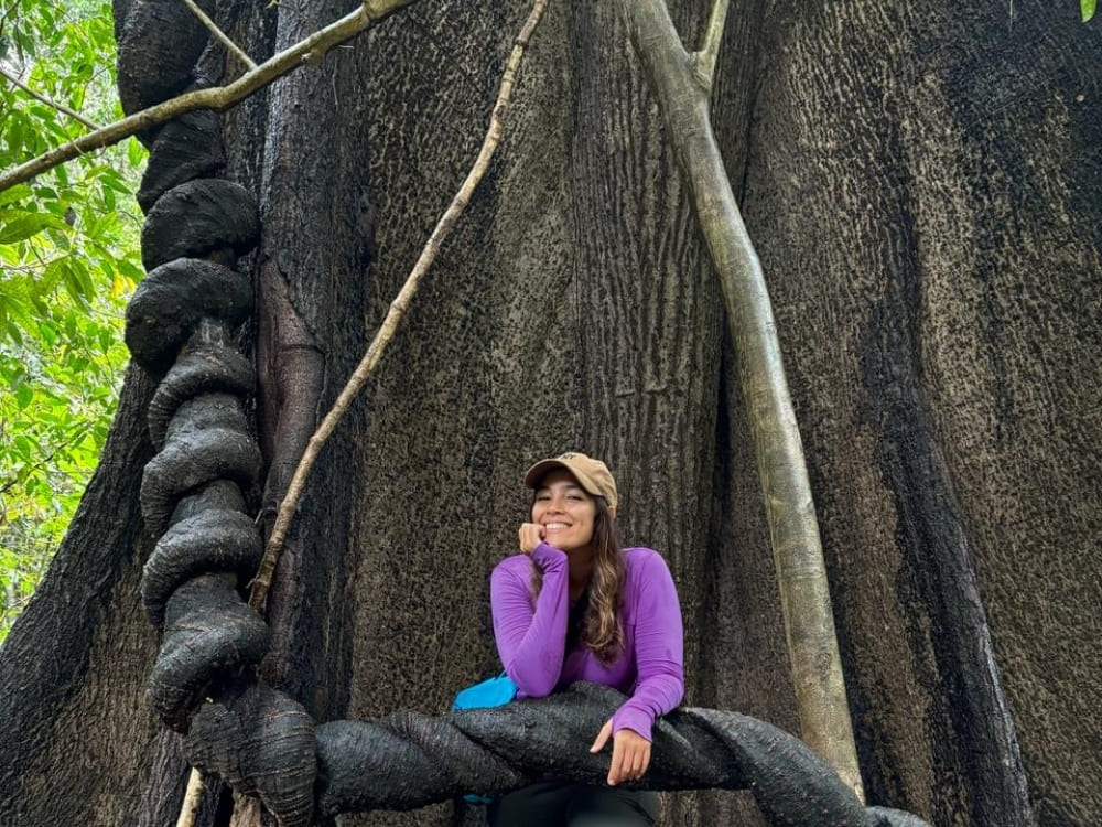 A smiling woman leans against thick, twisted vines at the base of a massive tree trunk in a tropical forest.