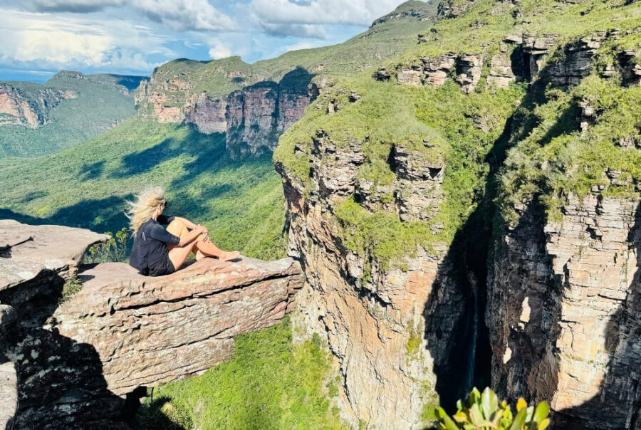 A blonde woman sits on a protruding rock ledge, looking out over a deep canyon and lush green valley under a sunny sky.